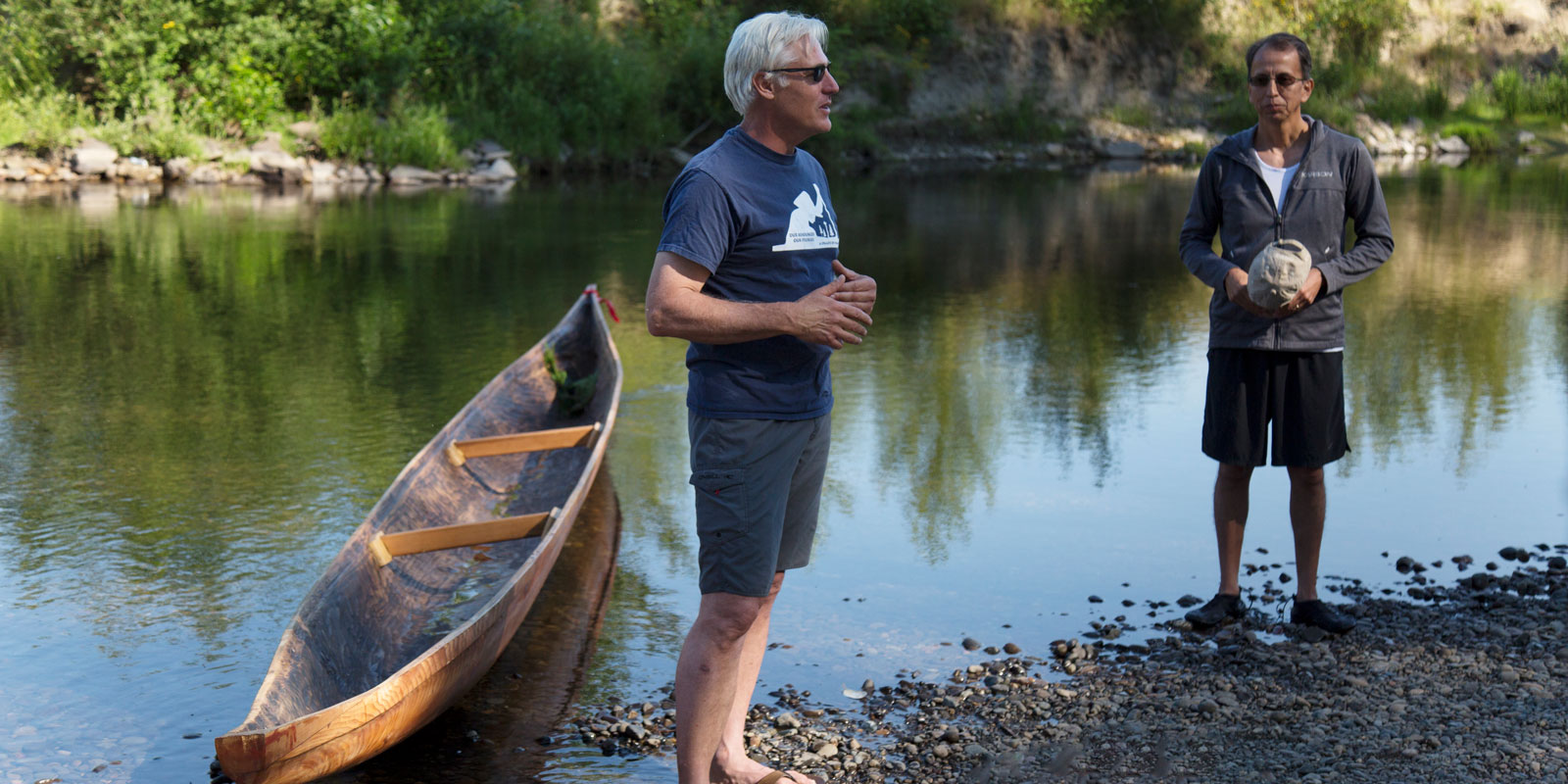 two men stand next to the canoe on the river bank