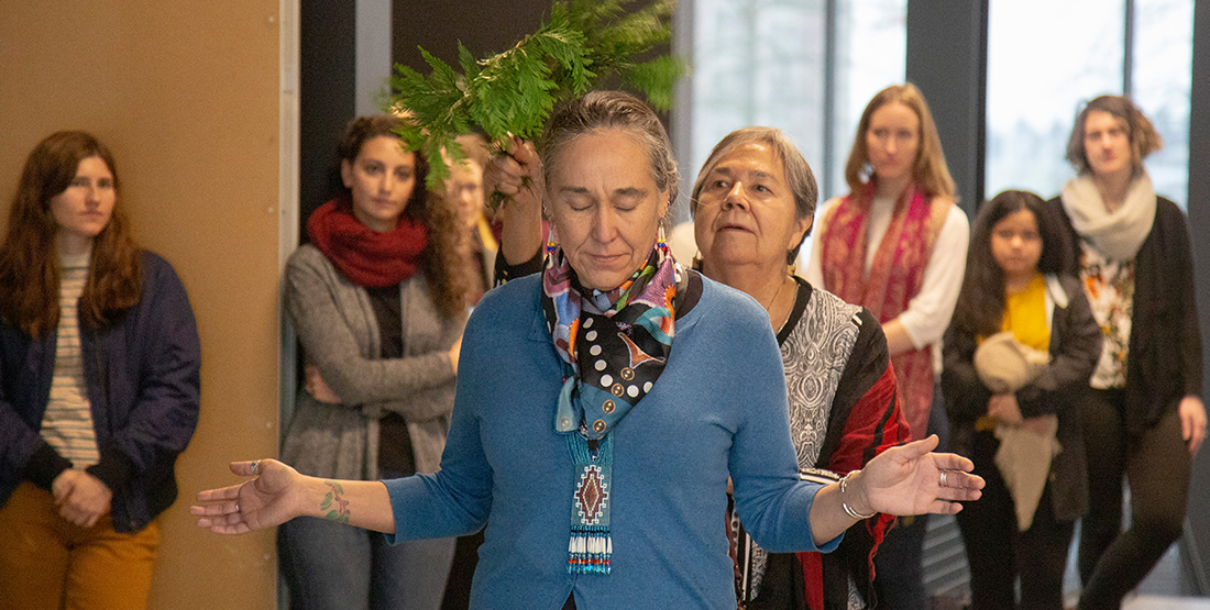 a woman stands with her hands out as she is brushed with cedar branches