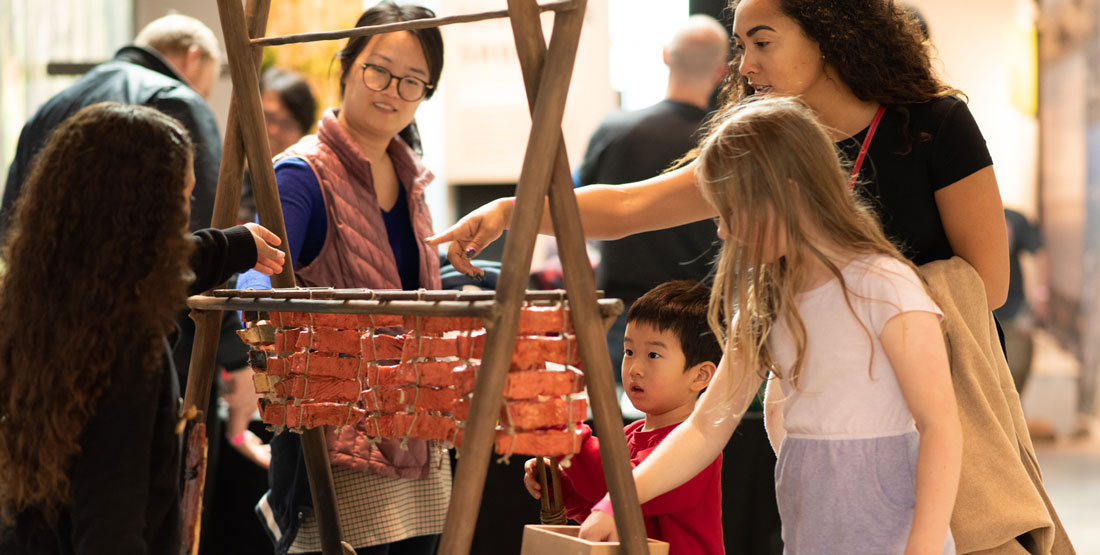 families touch an interactive exhibit depicting a salmon cooking over a fire pit