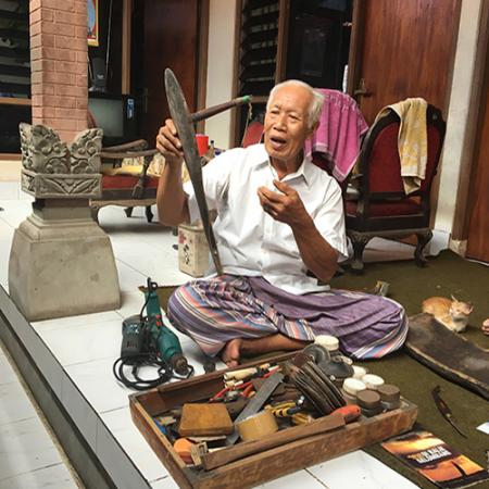 A man sits on a floor surrounded by fishing equipment and is holding a wooden tuna lure