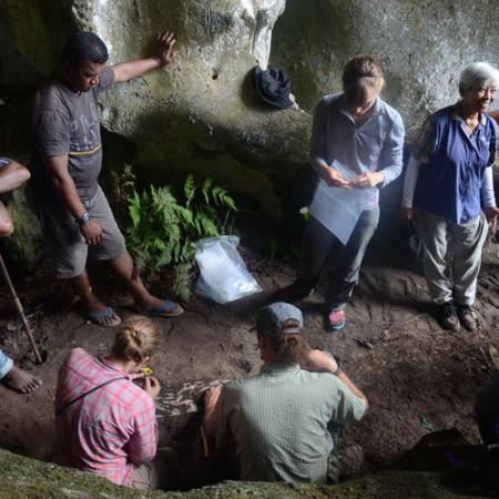 a group of people stand and watch as two researchers kneel on the ground and work