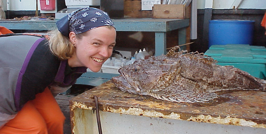 A woman smiles for the camera while posing with her face next to a large fish that had just been caught on a fishing vessel