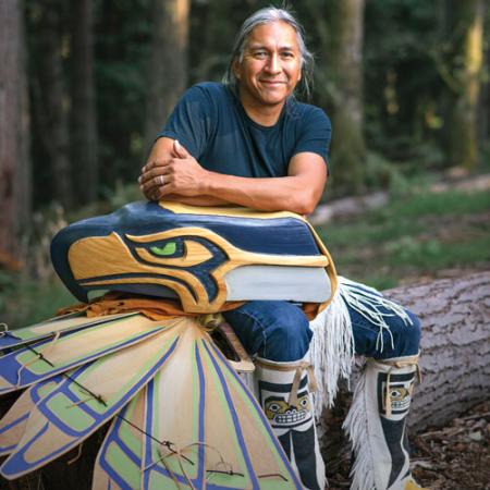 A man in a blue shirt sits with a mask that looks like the Seahawks logo and ceremonial clothes that look like wings