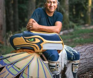 A man in a blue shirt sits with a mask that looks like the Seahawks logo and ceremonial clothes that look like wings