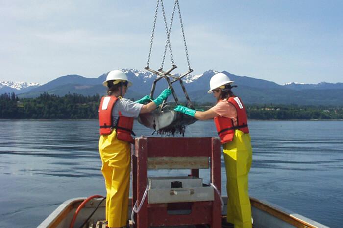 two people wear rain gear and hard hats while emptying sediment samples onto their boat