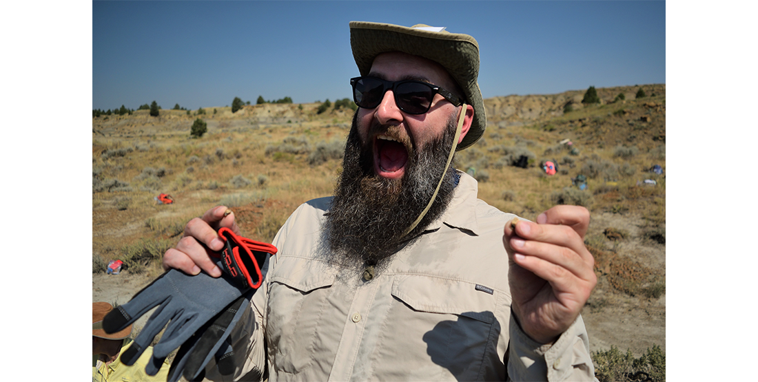 a person wearing a hat and sunglasses holds a microfossil in one hand